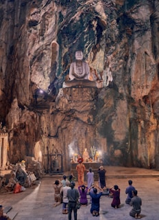A large statue of Buddha is carved into the rock face inside a cavernous temple space. Below, a group of people kneel on the ground, facing a lit altar adorned with offerings and incense. The atmosphere is serene and contemplative, with the natural rock formations towering above.