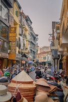 A bustling street scene with people walking and riding motorcycles past shops and buildings. The storefronts feature signs in various languages, and there are stacks of traditional conical hats in the foreground. The architecture is a mix of old and new styles, with narrow balconies and colorful facades.