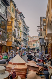A bustling street scene with people walking and riding motorcycles past shops and buildings. The storefronts feature signs in various languages, and there are stacks of traditional conical hats in the foreground. The architecture is a mix of old and new styles, with narrow balconies and colorful facades.