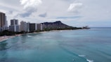 A coastal scene with a city skyline featuring tall buildings along a sandy beach. The turquoise ocean stretches out from the shoreline, with several surfers visible in the water. In the background, a prominent mountain rises under a partly cloudy sky.