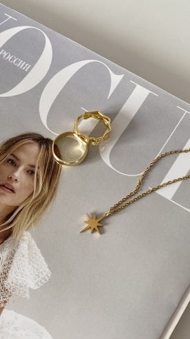 A fashion magazine featuring a model in a white lace dress. Resting on the magazine cover are two pieces of gold jewelry: a star-shaped pendant on a chain and two gold rings, one with an intricate design.