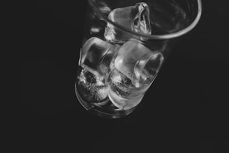 A transparent glass filled with ice cubes is positioned against a dark background. The ice cubes are stacked irregularly, with light reflecting off their smooth surfaces, creating a shiny and crisp texture.