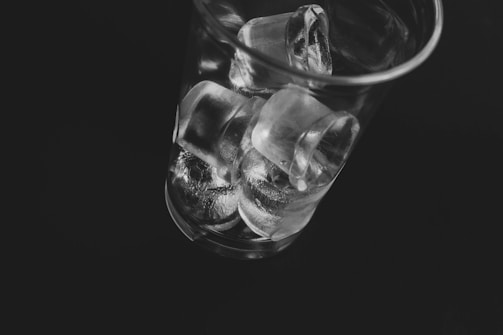 A transparent glass filled with ice cubes is positioned against a dark background. The ice cubes are stacked irregularly, with light reflecting off their smooth surfaces, creating a shiny and crisp texture.