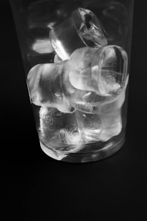 A clear glass filled with several ice cubes is positioned against a dark background. The ice cubes are transparent and stacked at varying angles within the glass, with surfaces showing subtle reflections and refractions.