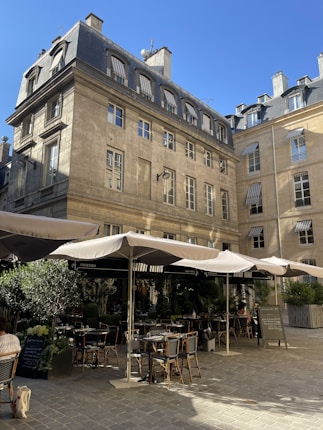Outdoor seating area of a caf&eacute; or restaurant with large beige umbrellas providing shade over tables and chairs. The establishment is located in an enclosed courtyard surrounded by classic European-style buildings with multiple windows and steep, slate rooftops. There are some green plants and potted trees around the seating area. The sky is clear and blue.