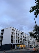 A modern multi-story apartment building with large windows and balconies. The structure is primarily composed of white and black materials. In the foreground, there is a street with a car driving by, and a few pedestrians walking along the sidewalk. Overhead, the sky is overcast, and a few trees partially frame the scene.