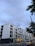 A modern multi-story apartment building with large windows and balconies. The structure is primarily composed of white and black materials. In the foreground, there is a street with a car driving by, and a few pedestrians walking along the sidewalk. Overhead, the sky is overcast, and a few trees partially frame the scene.
