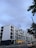 A modern multi-story apartment building with large windows and balconies. The structure is primarily composed of white and black materials. In the foreground, there is a street with a car driving by, and a few pedestrians walking along the sidewalk. Overhead, the sky is overcast, and a few trees partially frame the scene.