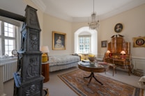 A vintage-style living room features ornate wooden furniture, including a round table and a detailed cabinet. A dark intricate stove is positioned to the left. The walls are adorned with classical portraits in gold frames. A chandelier hangs from the ceiling, adding elegance. The room has a cozy, historic vibe with a patterned carpet and light streaming through the window.