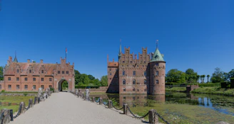 A medieval-style castle with red brick walls and multiple turrets sits alongside a tranquil moat. A stone pathway leads to an arched entrance, surrounded by lush green vegetation under a clear blue sky.