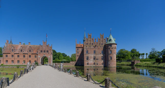 A medieval-style castle with red brick walls and multiple turrets sits alongside a tranquil moat. A stone pathway leads to an arched entrance, surrounded by lush green vegetation under a clear blue sky.