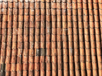 A repetitive pattern of weathered, red-brown roof tiles, arranged in neat rows, with visible wear and some areas of black discoloration and moss growth.