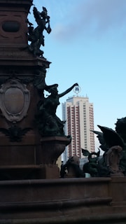A detailed, ornate monument with statues is depicted in the foreground against the backdrop of a modern high-rise building. The statues feature intricate sculpting, including figures of angels and a central figure holding a metallic object. The high-rise in the background is a tall, sleek building with a distinct 'M' logo at the top.