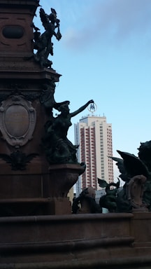 A detailed, ornate monument with statues is depicted in the foreground against the backdrop of a modern high-rise building. The statues feature intricate sculpting, including figures of angels and a central figure holding a metallic object. The high-rise in the background is a tall, sleek building with a distinct 'M' logo at the top.