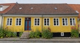 A charming, single-story house with a vibrant yellow façade and a distinctive red-tiled roof. The house features a centrally located ornate blue and red door, flanked by multiple white-framed windows. Lush green plants and blooming pink flowers adorn the front, adding a touch of nature to the setting. The cobblestone path in front enhances the quaint and rustic appeal of the house.