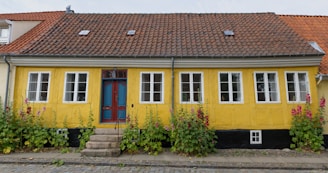 A charming, single-story house with a vibrant yellow façade and a distinctive red-tiled roof. The house features a centrally located ornate blue and red door, flanked by multiple white-framed windows. Lush green plants and blooming pink flowers adorn the front, adding a touch of nature to the setting. The cobblestone path in front enhances the quaint and rustic appeal of the house.