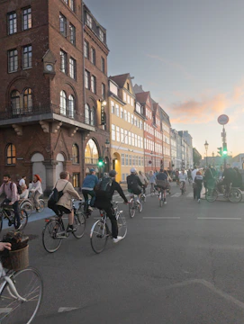 A group of cyclists riding stylish commuter bikes through a bustling city street near a bike business storefront.