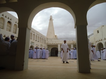 A group of people dressed in white robes and head coverings gather in a courtyard with arches and a central tall tower. The architecture features ornate geometric patterns on the windows and an overcast sky above.
