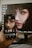 A hand is holding a smartphone displaying the same digital artwork shown on a computer monitor in the background. The artwork features a close-up of a woman's face with striking, stylized makeup around the eyes, giving the image a fantasy or abstract vibe. The smartphone and the monitor share the same image, showing synchrony.