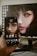 A hand is holding a smartphone displaying the same digital artwork shown on a computer monitor in the background. The artwork features a close-up of a woman's face with striking, stylized makeup around the eyes, giving the image a fantasy or abstract vibe. The smartphone and the monitor share the same image, showing synchrony.