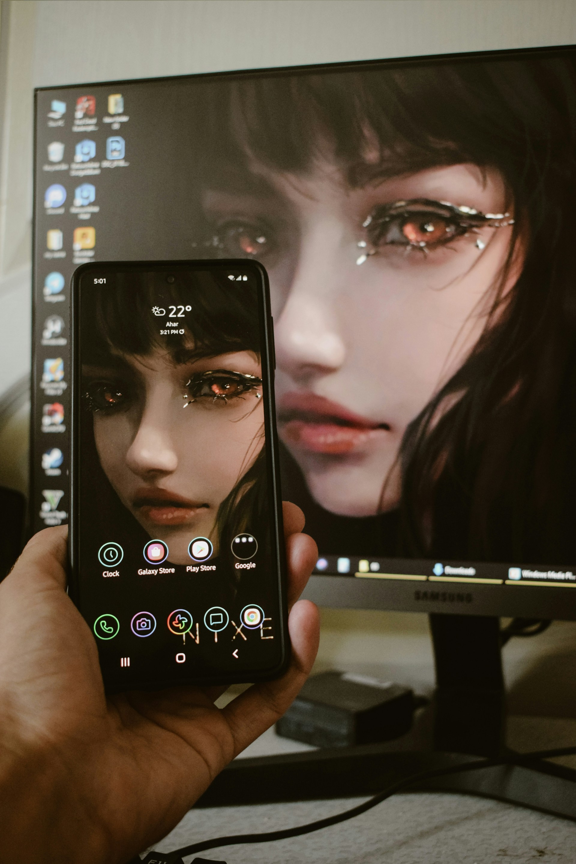 A hand is holding a smartphone displaying the same digital artwork shown on a computer monitor in the background. The artwork features a close-up of a woman's face with striking, stylized makeup around the eyes, giving the image a fantasy or abstract vibe. The smartphone and the monitor share the same image, showing synchrony.