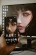A hand is holding a smartphone displaying the same digital artwork shown on a computer monitor in the background. The artwork features a close-up of a woman's face with striking, stylized makeup around the eyes, giving the image a fantasy or abstract vibe. The smartphone and the monitor share the same image, showing synchrony.