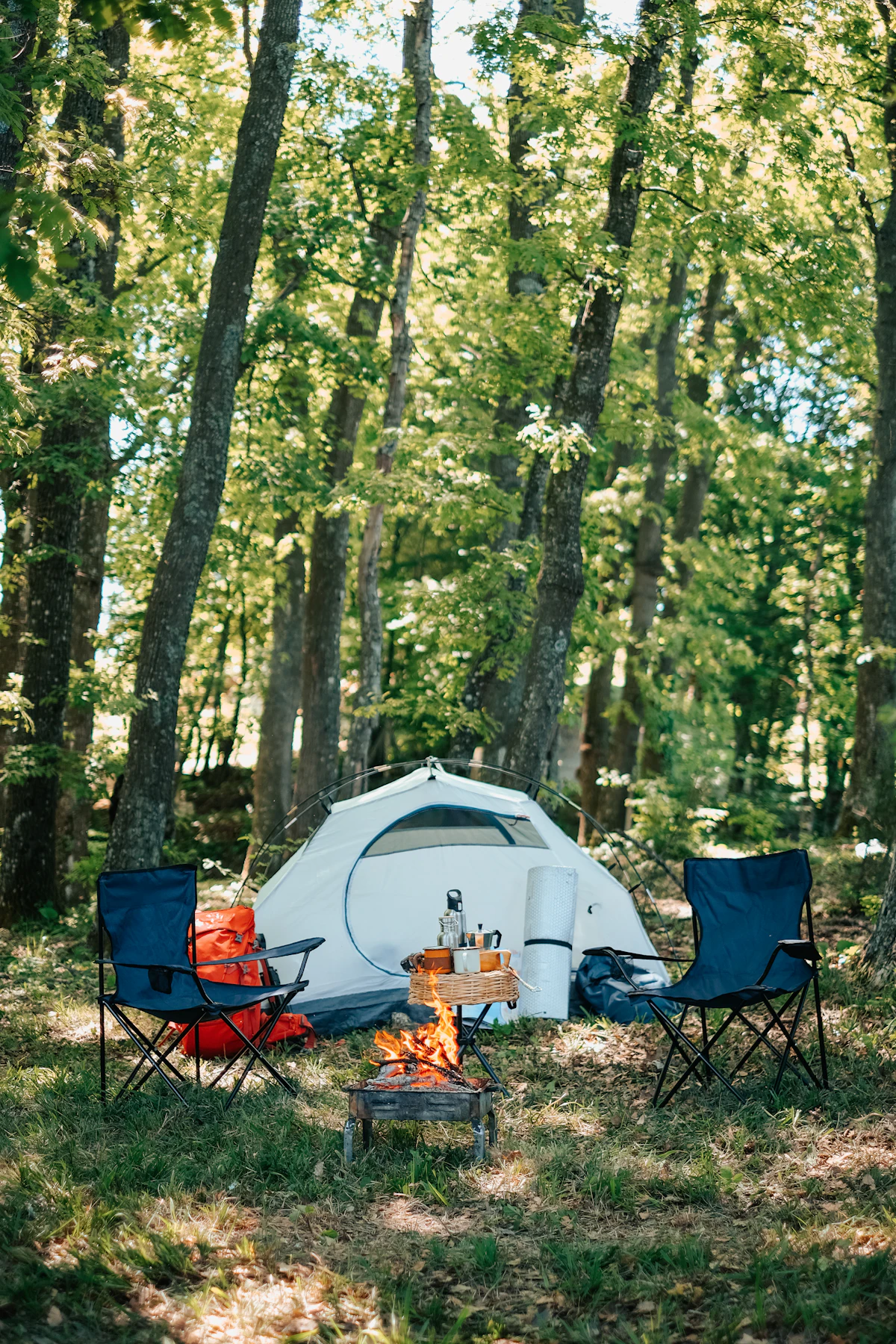 A complete outdoor campsite with a tent, portable chairs, camping gear, and a small campfire in a forest