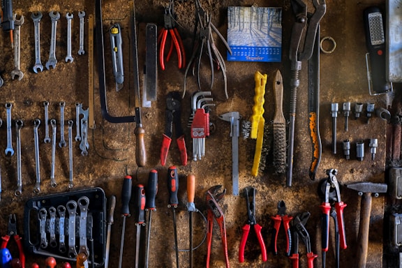 A rustic workshop wall is covered with neatly arranged tools, including wrenches, pliers, screwdrivers, and saws. A small calendar hangs on the wall, adding to the organized chaos. The tools vary in color and size, indicating their different uses.