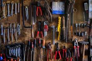 A rustic workshop wall is covered with neatly arranged tools, including wrenches, pliers, screwdrivers, and saws. A small calendar hangs on the wall, adding to the organized chaos. The tools vary in color and size, indicating their different uses.