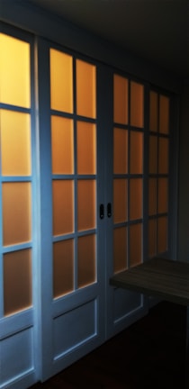 A set of sliding glass doors with a wooden frame, partially illuminated by a warm light from behind. The panels are arranged in a grid pattern, casting a soft glow through the translucent glass.