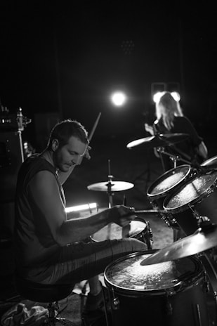 A drummer is immersed in playing a drum set under dim lighting. Another musician, possibly playing the guitar, is seen in the background with bright stage lights illuminating from behind. The atmosphere reflects a live music performance, with focus on the concentration and energy of the drummer.