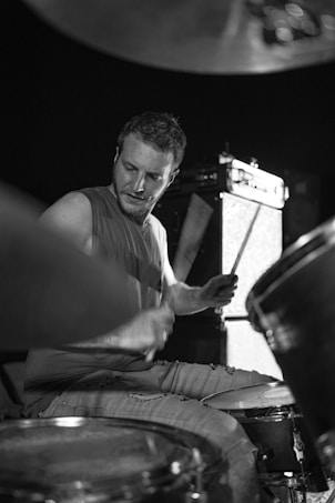 A drummer captured in mid-performance, intensely focused on playing a drum set. The image is in black and white, highlighting the contrast and dynamic movement of the drumsticks. The background features an amplifier, suggesting a live music setting.
