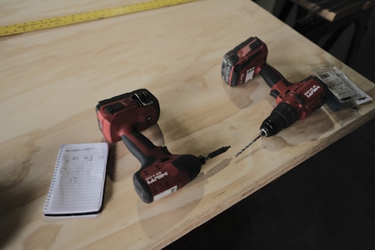 Two red cordless power drills are placed on a wooden surface along with a small spiral notebook containing handwritten numbers. A yellow measuring tape stretches across the table in the background.