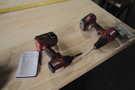 Two red cordless power drills are placed on a wooden surface along with a small spiral notebook containing handwritten numbers. A yellow measuring tape stretches across the table in the background.
