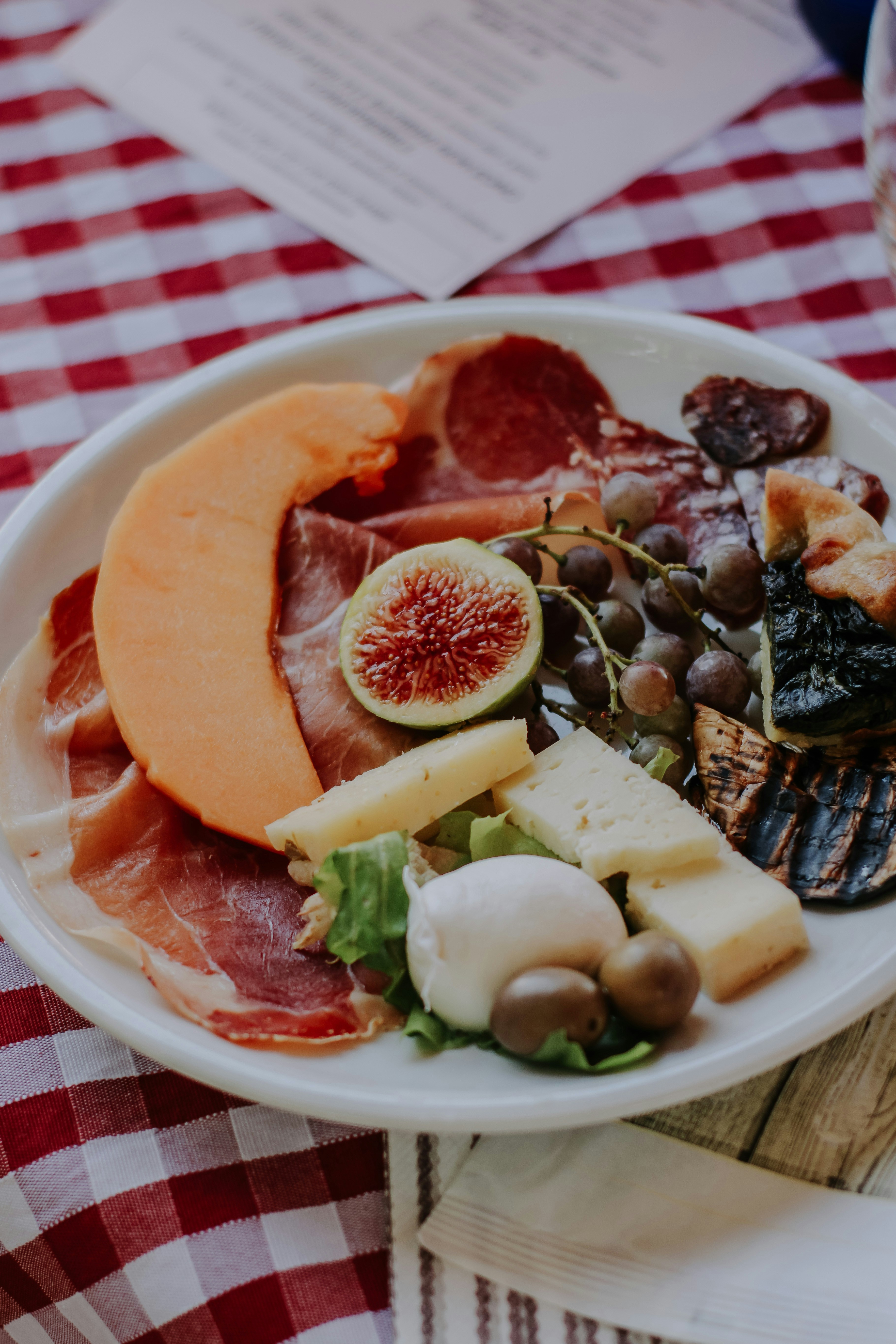 White plate with arranged cured meats, cheeses, figs, and vegetables on checkered tablecloth