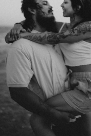 A monochrome image depicting a close-up of a couple in an intimate pose. The man with a beard is holding a woman who has tattoos on her arms and is wearing a tank top and shorts. Their expressions suggest a moment of affection.