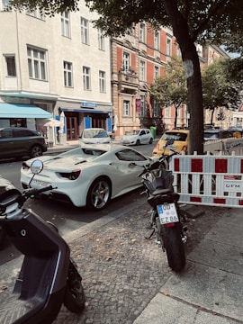 A street scene featuring a white sports car parked on the side of the road, with a motorcycle and scooter parked nearby. The street is lined with trees and adjacent to an older styled building with white and red brick facade. There are other vehicles parked along the street and a shop with a blue sign is visible in the background.
