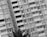 A black and white photograph featuring a tall residential building with numerous balconies and windows. The image is captured at a slight angle, adding a dynamic perspective. A large palm tree is visible in the foreground, its leaves reaching toward the building.