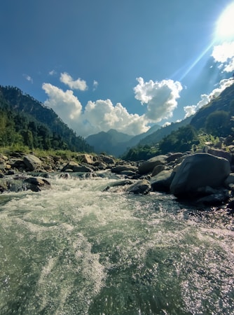 A clear river flows energetically over rocks and pebbles, nestled between lush green mountains under a vast blue sky. Fluffy white clouds float above, with sunlight streaming through, casting highlights on the water and surrounding landscape.