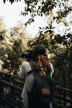 A person wearing a cap and carrying a camera is walking on a wooden pathway surrounded by lush green trees. The individual is equipped with a large backpack, suggesting they are on a hiking or nature photography expedition. The setting appears tranquil and shaded by the canopy of leaves above.