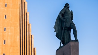 A statue of a heroic figure wearing a cloak and standing in a confident pose on a pedestal. To the left, there is a tall building with a unique architectural design featuring a series of vertical ridges and small, evenly spaced windows. The sky is clear and blue.