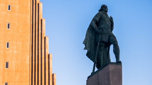 A statue of a heroic figure wearing a cloak and standing in a confident pose on a pedestal. To the left, there is a tall building with a unique architectural design featuring a series of vertical ridges and small, evenly spaced windows. The sky is clear and blue.