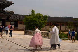 People are walking in a traditional Korean architectural setting, with a focus on individuals wearing traditional Korean clothing, hanbok. The background features historical-style buildings and a vibrant green tree in the courtyard.