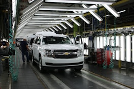 A vehicle assembly line inside an automotive factory, featuring a white SUV at the forefront. Bright overhead lights illuminate the work area, with several workers engaged in the production process. The SUV is stationed on a conveyor belt, with nearby tools and equipment supporting the assembly work.