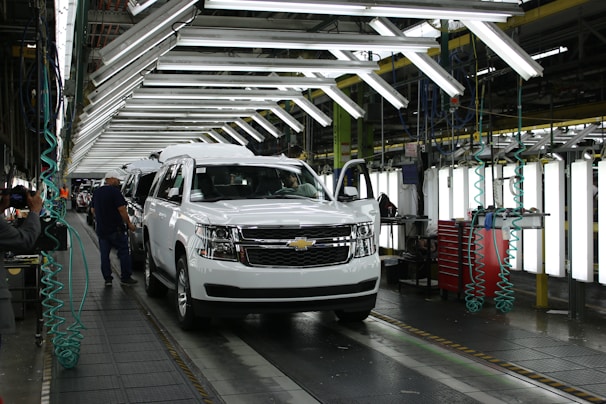 A vehicle assembly line inside an automotive factory, featuring a white SUV at the forefront. Bright overhead lights illuminate the work area, with several workers engaged in the production process. The SUV is stationed on a conveyor belt, with nearby tools and equipment supporting the assembly work.