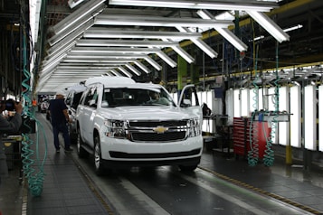 A vehicle assembly line inside an automotive factory, featuring a white SUV at the forefront. Bright overhead lights illuminate the work area, with several workers engaged in the production process. The SUV is stationed on a conveyor belt, with nearby tools and equipment supporting the assembly work.