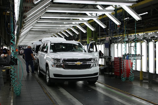 A vehicle assembly line inside an automotive factory, featuring a white SUV at the forefront. Bright overhead lights illuminate the work area, with several workers engaged in the production process. The SUV is stationed on a conveyor belt, with nearby tools and equipment supporting the assembly work.