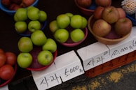 Several baskets filled with green and red apples are placed on a table along with visible price tags written in Korean. The apples are neatly arranged, and the setting suggests an outdoor market environment.