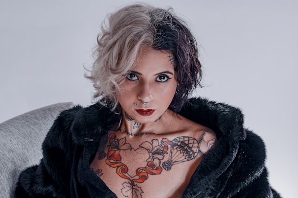 A person with a striking half-blonde, half-dark hairstyle, wearing dark eye makeup and dark red lipstick. Their chest displays a colorful tattoo with various designs. They are dressed in a black fur coat and have facial piercings.