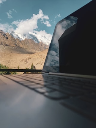 A partially open laptop is placed on a table, reflecting a stunning view of snow-capped mountains. The background features a clear blue sky with a few clouds and a rugged mountain range. The focus on the laptop blurs the natural scenery slightly, emphasizing the reflection on the laptop screen.